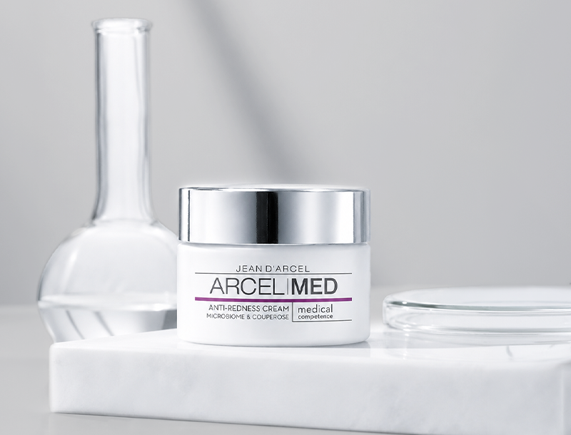 Tiegel der Jean D'Arcel ARCELMED Anti-Redness Cream auf weißem Marmorsockel neben einem Glasgefäß in heller, minimalistischer Umgebung.